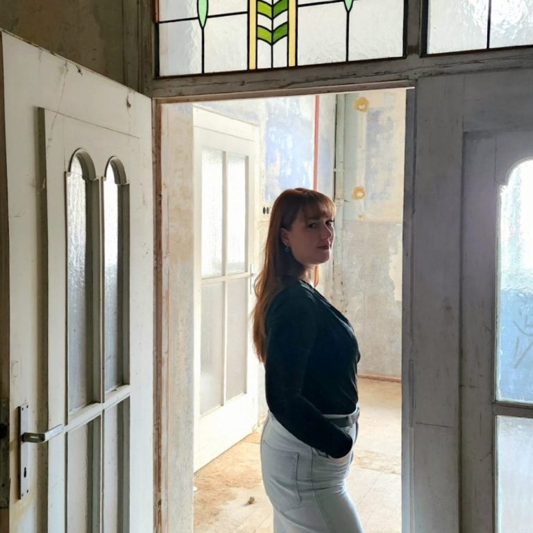 Die Galeristin steht in einem hellen Raum mit Flügeltür und Buntglasfenster im Jugendstil. Die Tür verbindet die Galerieräume miteinander.